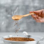 A close-up of a hand pouring flaxseeds into a bowl using a wooden spoon.